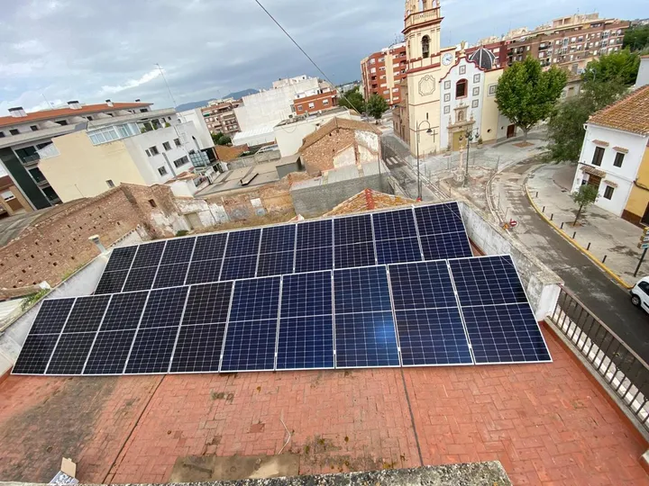Campo de paneles solares montado sobre la cubierta plana de un edificio, con el casco urbano y el campanario al fondo